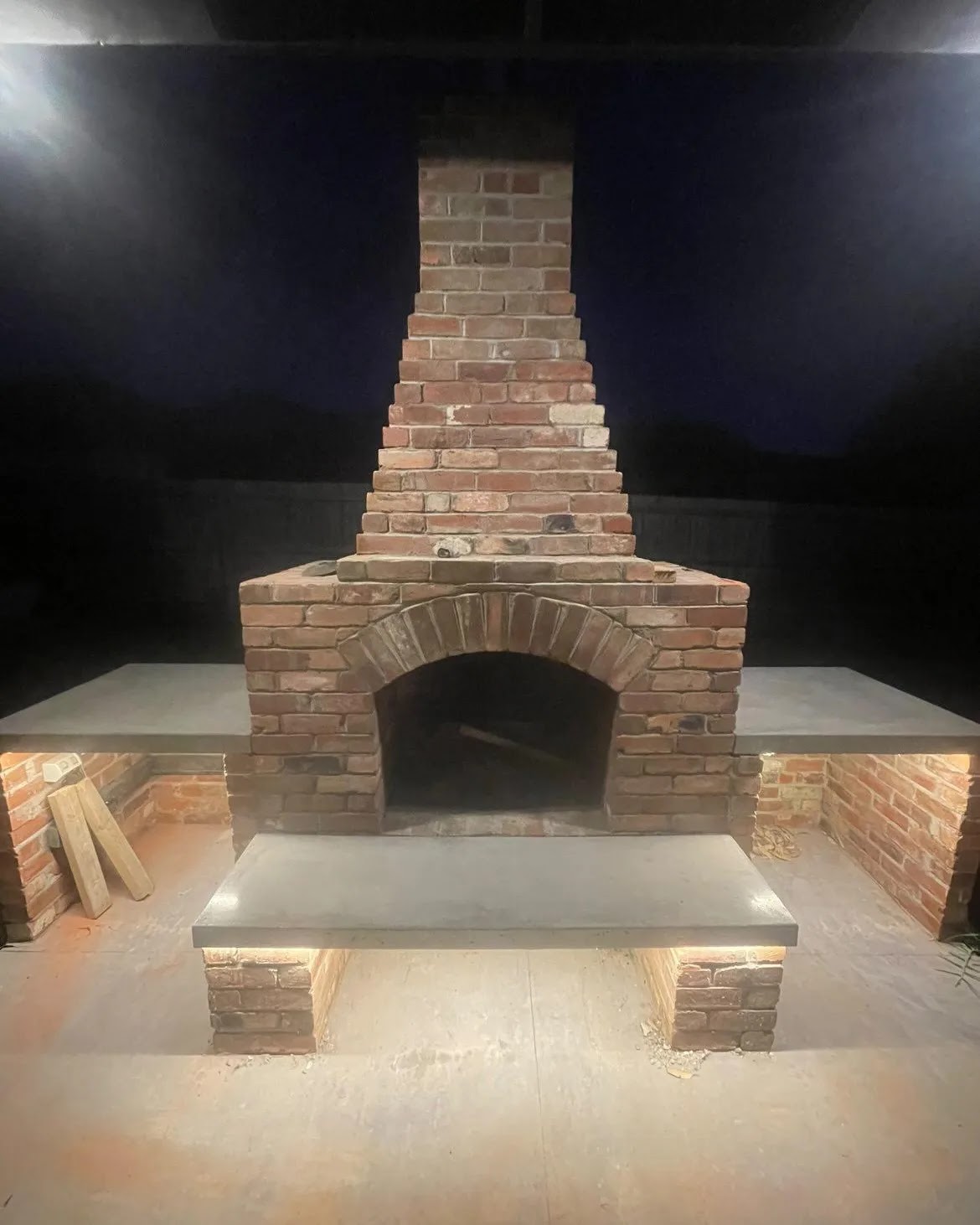Recycled brick outdoor fireplace at night with a concrete hearth bench and warm lighting underneath