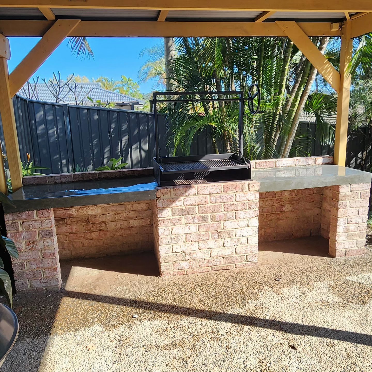 Outdoor BBQ kitchen with a red brick base and dark concrete bench top under a timber pergola
